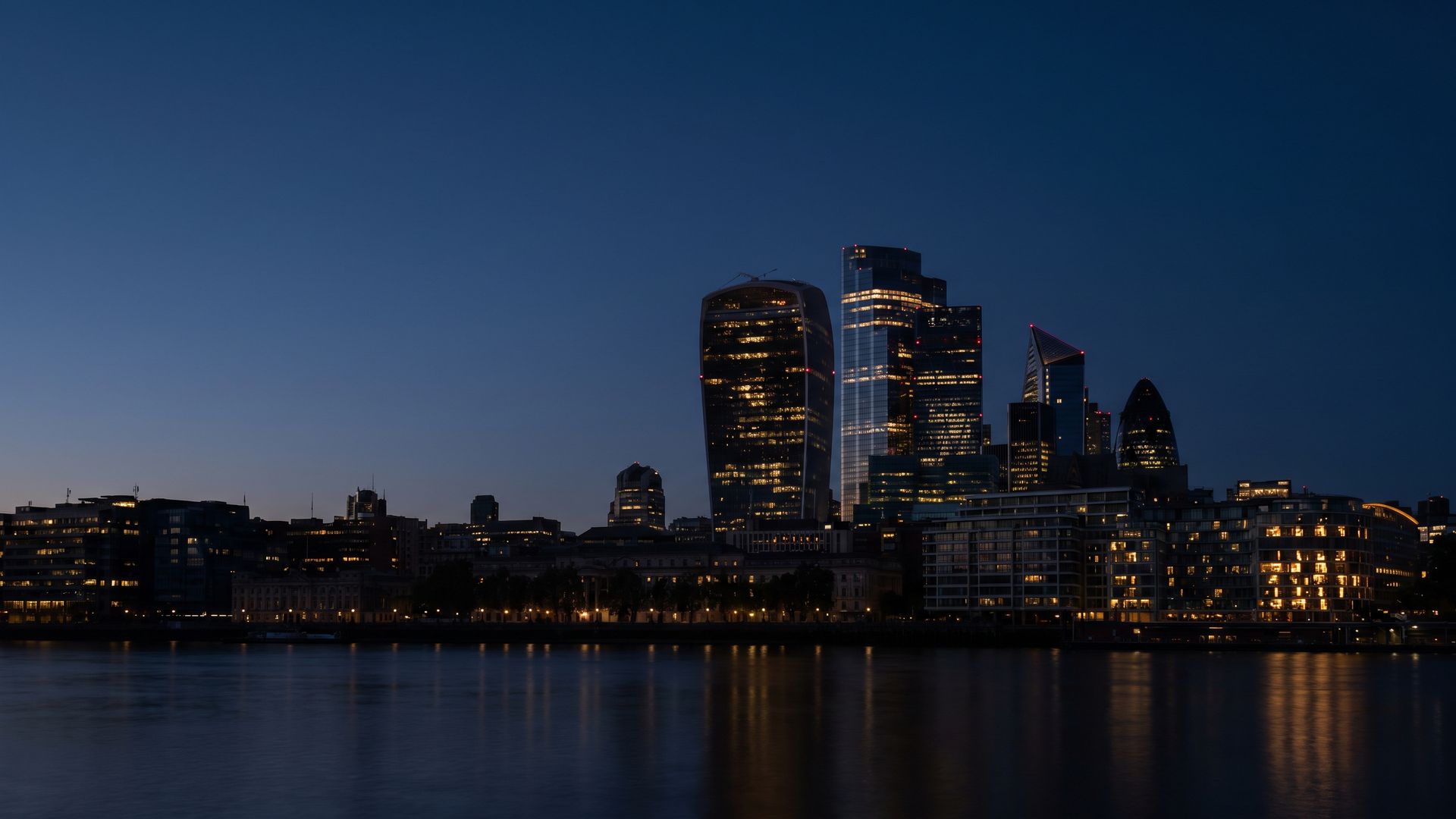 The City of London financial district at dusk, seen across the river, its towers lit against a deep blue sky.