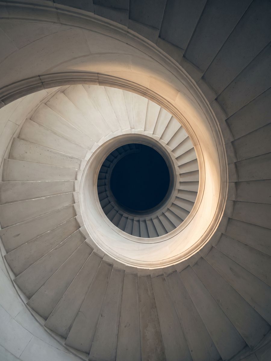 The underside of a stone spiral staircase curving up into shadow, one edge caught by daylight.